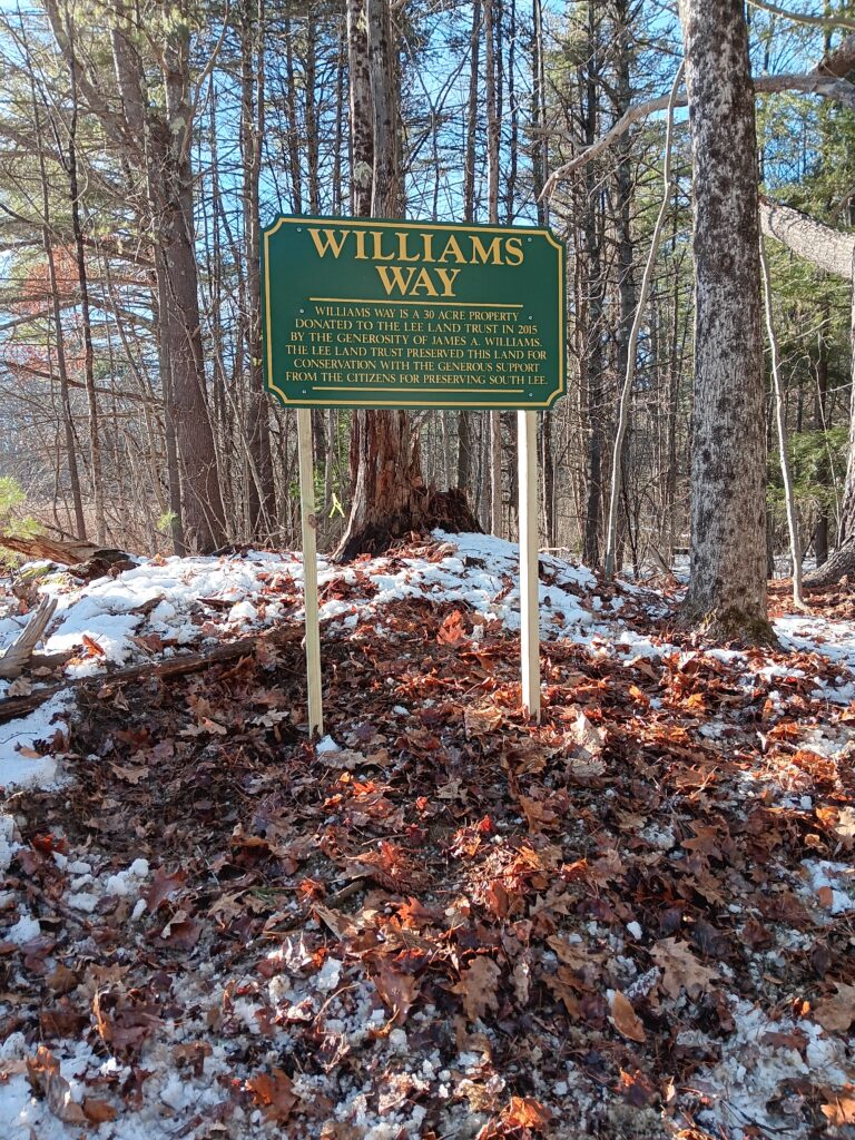 A green sign with gold lettering which says: Williams Way is a 30 acre property donated to the Lee Land Trust in 2015 by the generosity of james A. Williams. The Lee Land Trust preserved this land for conservation with the generous support from the Citizens for Preserving South Lee.